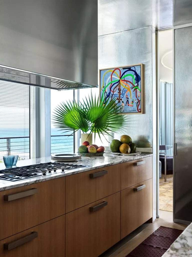 a kitchen has wood cabinets with a marble countertop and built in stovetop, titanium walls with a colorful abstract painting, terra cotta floor mats, windows face the sea