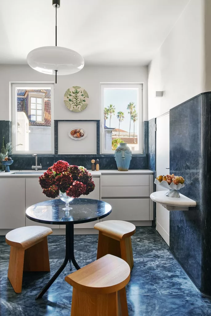 a kitchen floor, backsplash, partial wall, and a round table are a blue marble, three stools are oak, two windows are above white cabinets and sink, a floating shelf has a fruit bowl, a spheroid pendant above table