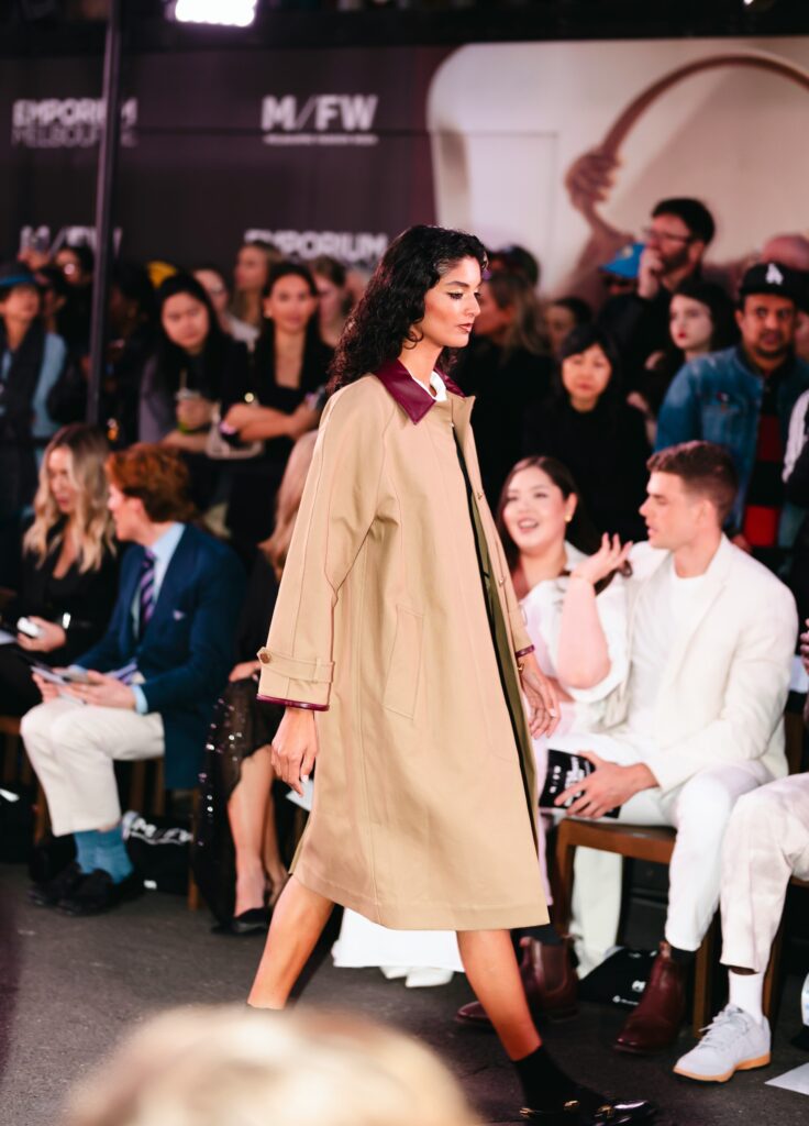 Personne défilant sur un podium, vêtue d’un long manteau beige agrémenté de détails bordeaux.