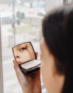 Gros plan d'une femme appliquant du maquillage pour les yeux avec un miroir de poche près d'une fenêtre.
