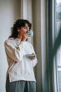 femme blanche avec des cheveux bruns ondulés qui boit son café en face d'une fenêtre