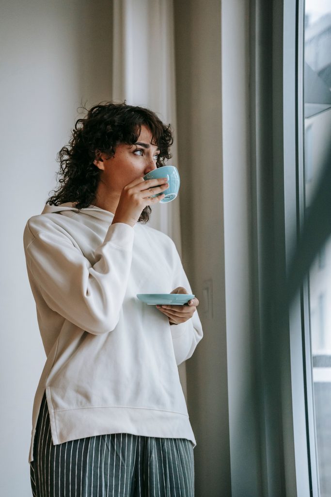 femme blanche avec des cheveux bruns ondulés qui boit son café en face d'une fenêtre