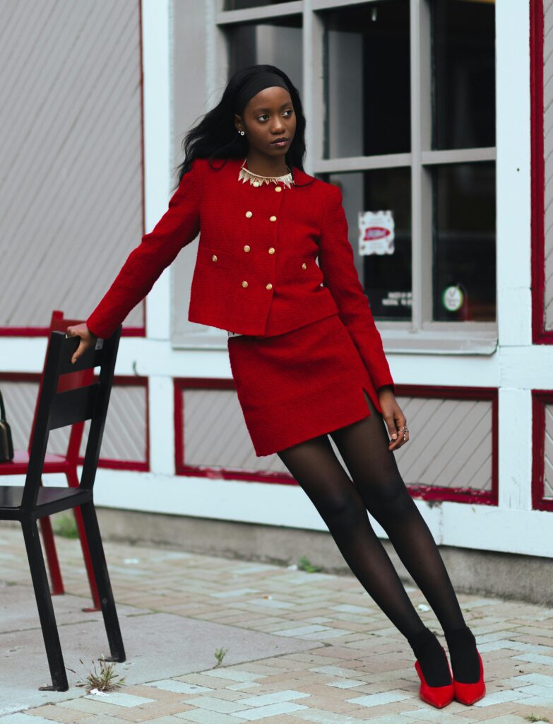 Personne vêtue d’un ensemble rouge composé d’un blazer et d’une jupe, assorti à des talons rouges. Elle se tient à une chaise.